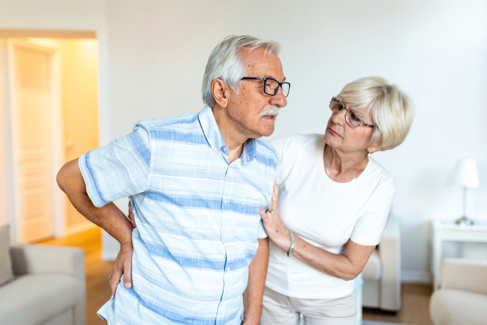 An elderly man with grey hair and glasses, holding his lower back in pain, supported by a concerned elderly woman