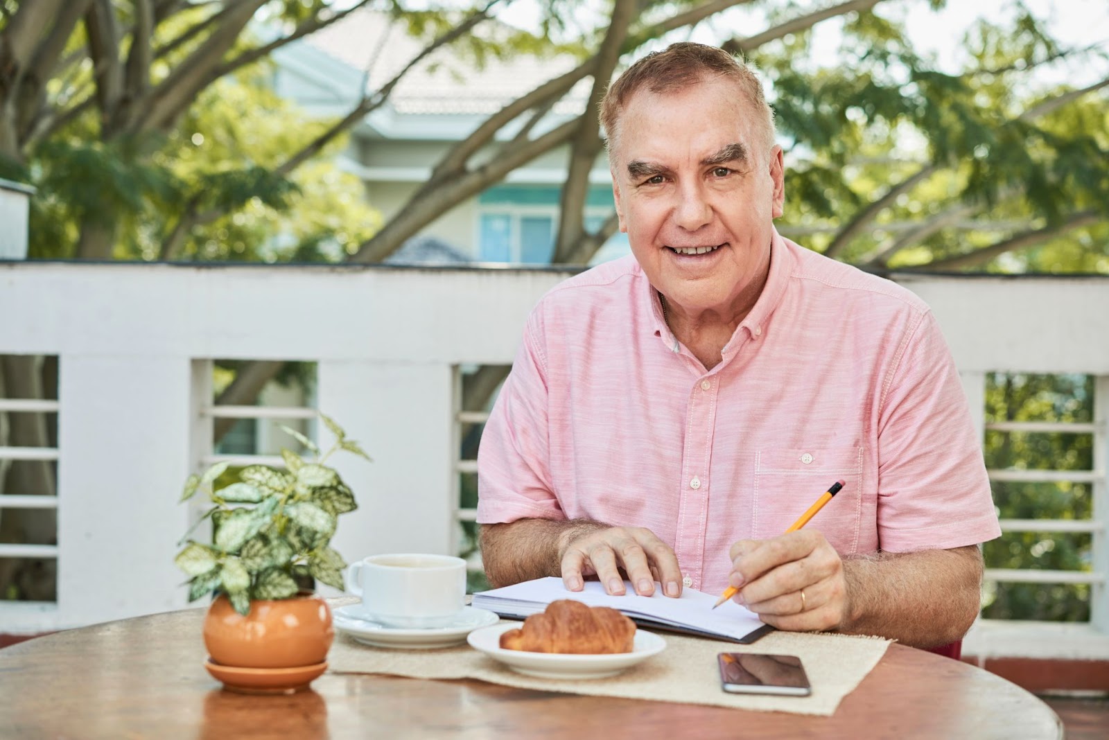 Older man writing down meal planning notes outdoors with fresh fruit and breakfast items on the table